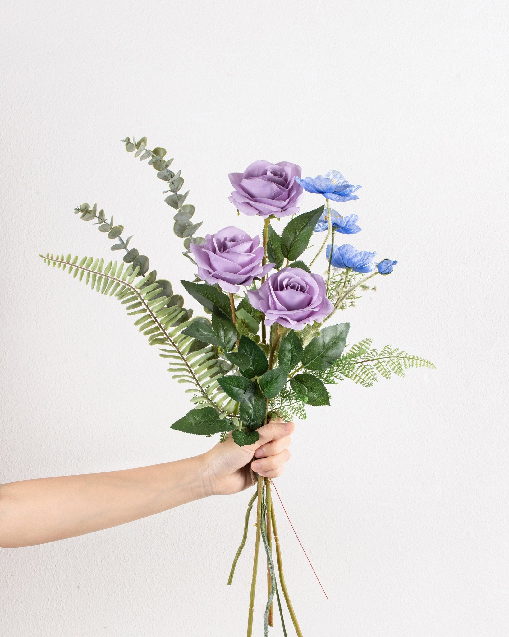 Hand holding a bouquet of artificial purple roses and blue poppies with green foliage against a white textured background.