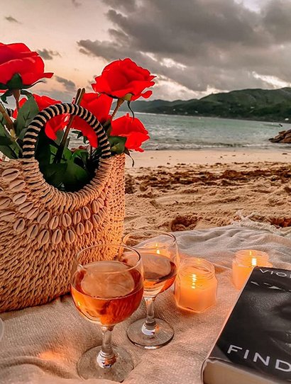 Artificial burgundy roses in a woven bag on a beach picnic with wine glasses and candles