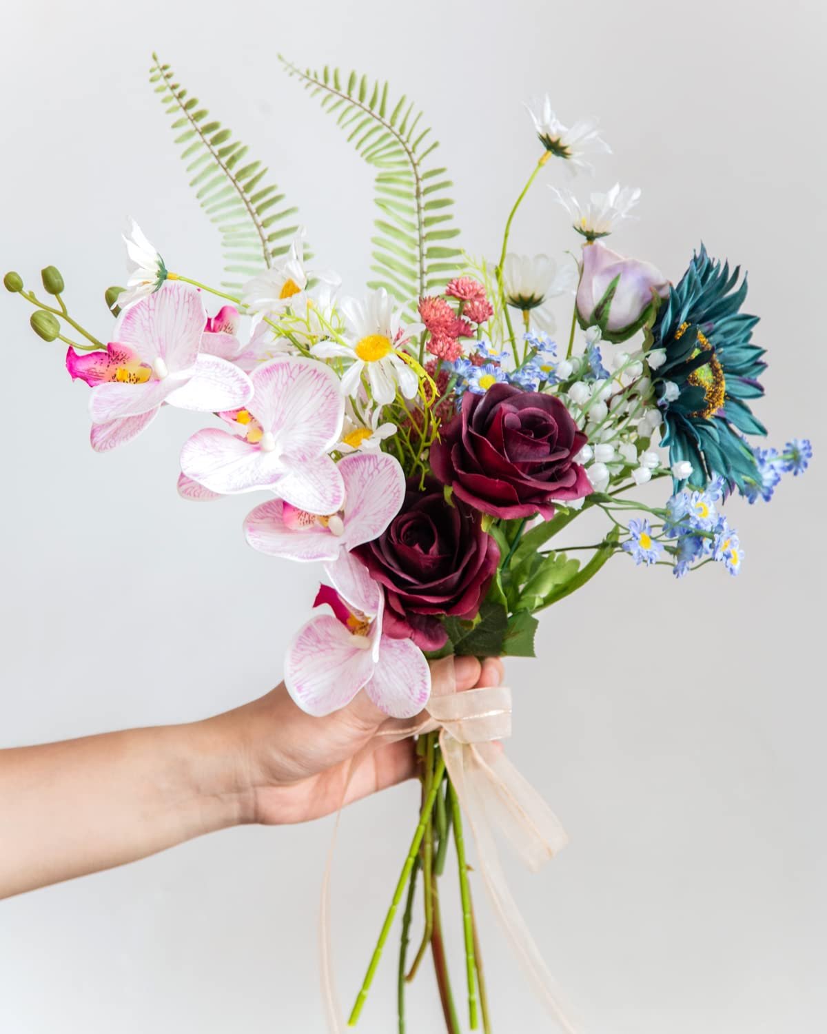 Hand holding a bouquet of artificial burgundy roses, pink orchids, and blue flowers with green ferns.