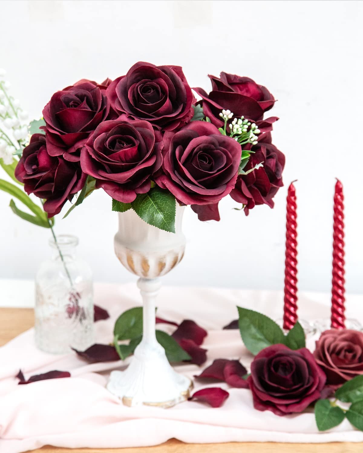 Burgundy silk roses arranged in a decorative vase with scattered petals and red candles