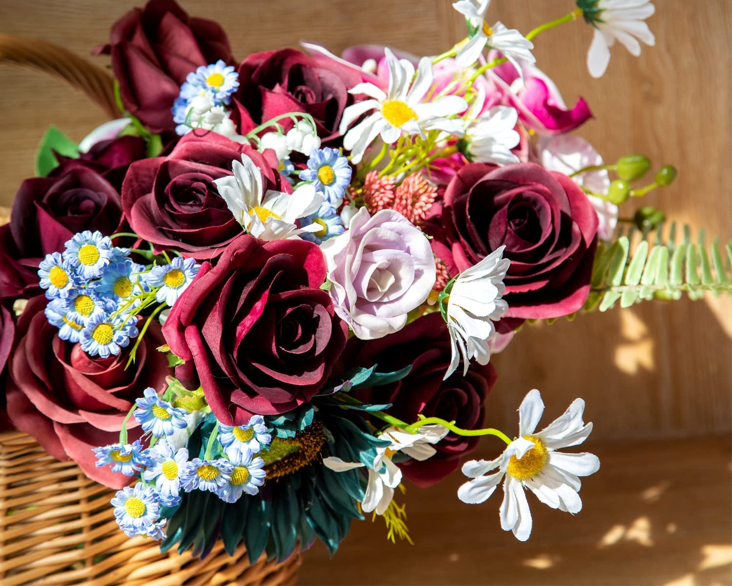 Burgundy silk rose bouquet with daisies and other flowers in a wicker basket on a wooden surface.