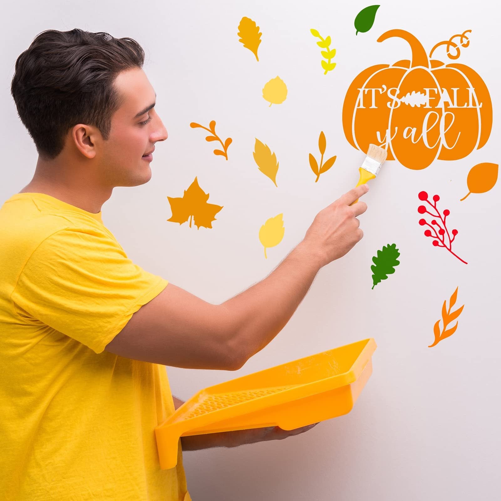 Man in yellow shirt painting autumn leaf and pumpkin stencils on a white wall with a brush and paint tray