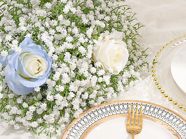 Close-up of artificial baby's breath with two roses, one blue and one white, on a table setting with a gold-rimmed plate and fork.