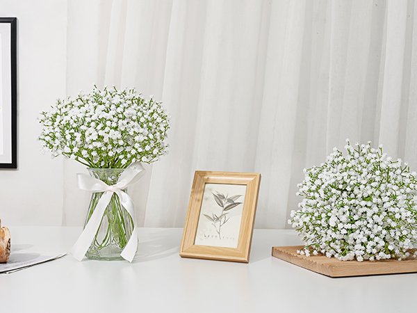 Two bouquets of white artificial baby's breath flowers, one in a glass vase with a ribbon, the other on a wooden stand, with a picture frame nearby.