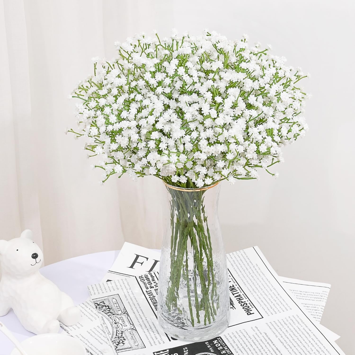 A bouquet of white artificial baby's breath flowers in a clear glass vase on a table with a white bear figurine and newspapers.