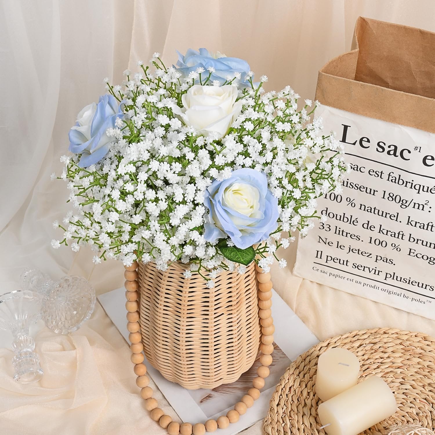 Artificial baby's breath and roses in a woven basket with wooden beads, next to candles and a paper bag.