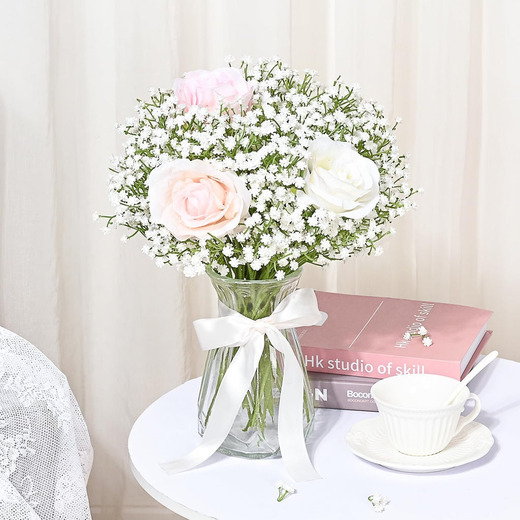 Bouquet of artificial baby's breath with pink and white roses in a glass vase, tied with a white ribbon, on a table with books and a teacup.
