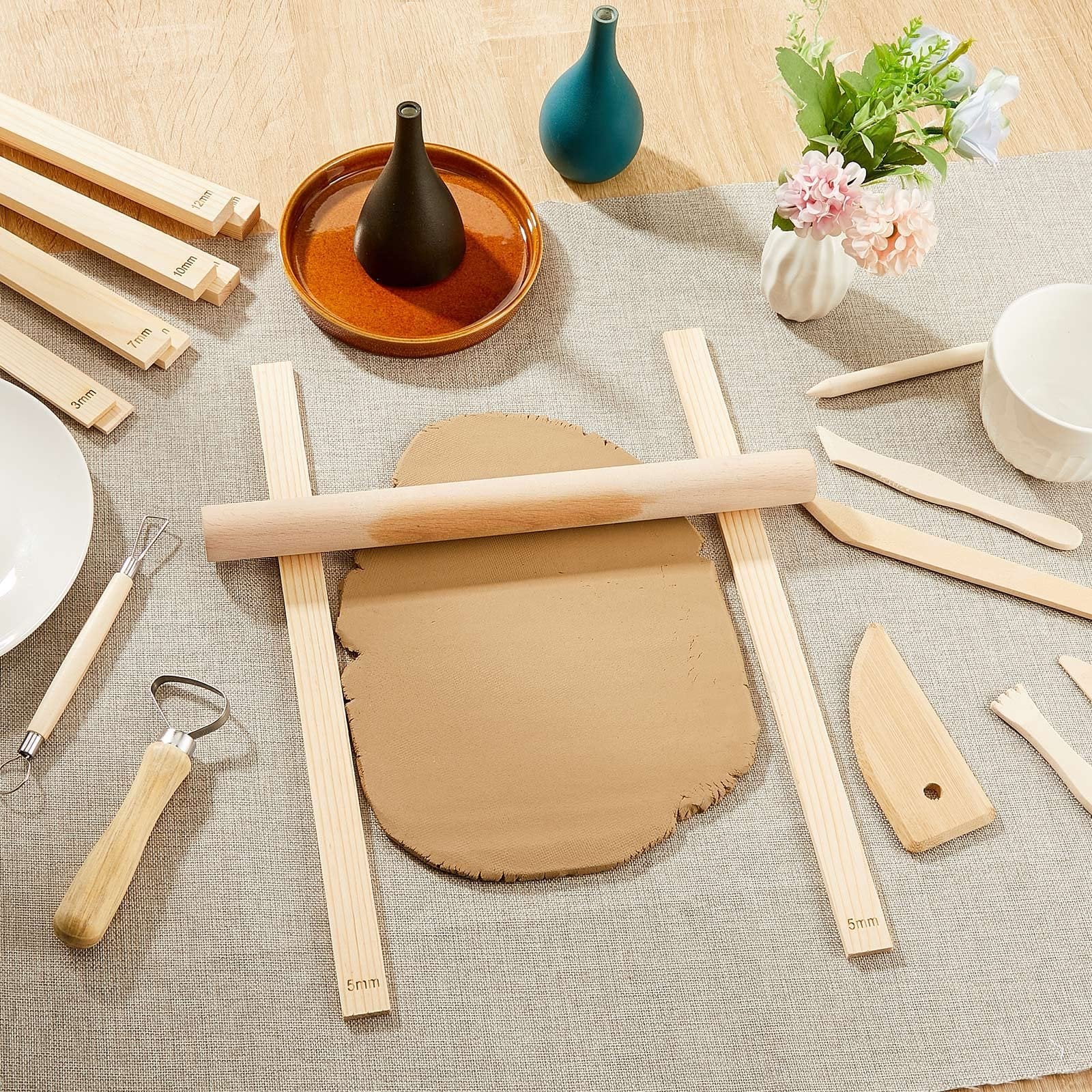Clay rolling pin guides and tools arranged on a table with clay and decorative vases.