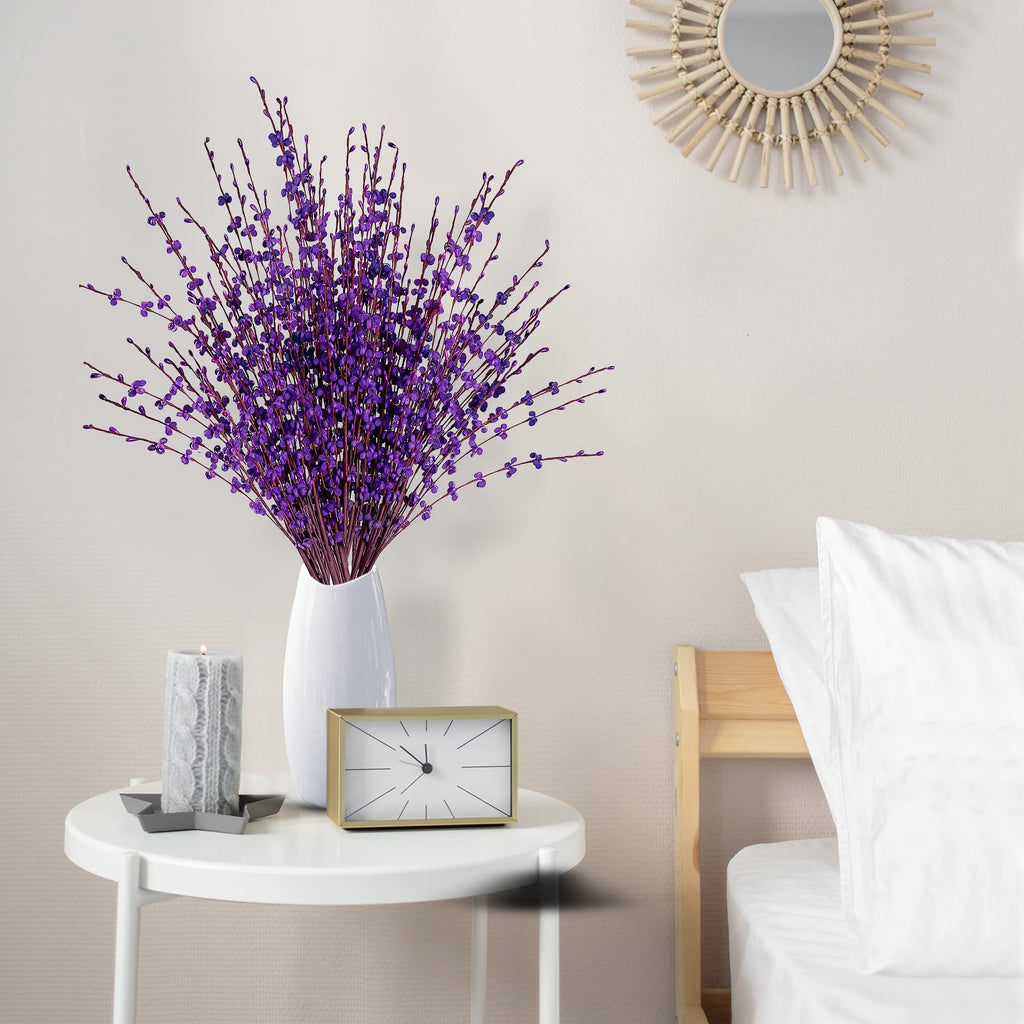 Bouquet of dark purple artificial jasmine flowers in a white vase on a side table with a candle and clock, next to a bed.