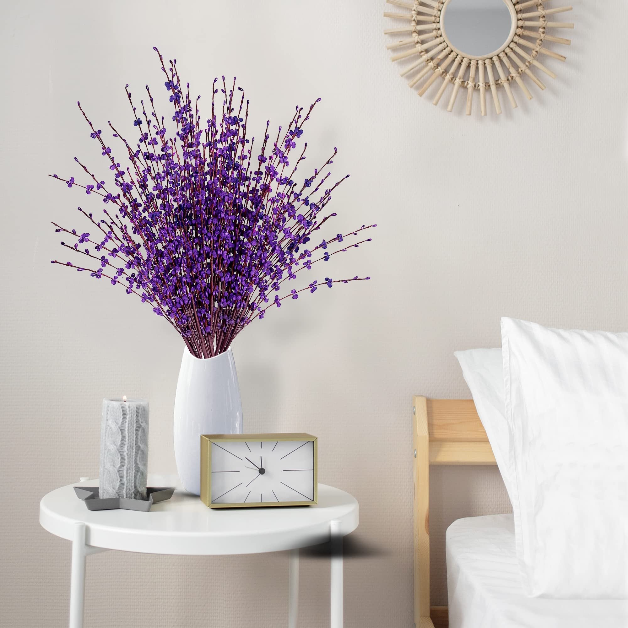 Bouquet of dark purple artificial jasmine flowers in a white vase on a side table with a candle and clock, next to a bed.