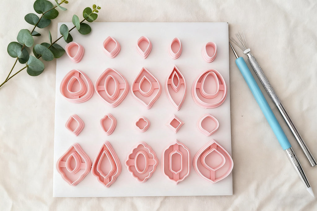 Set of pink polymer clay earring cutters in various shapes arranged on a white board with eucalyptus leaves and sculpting tools.