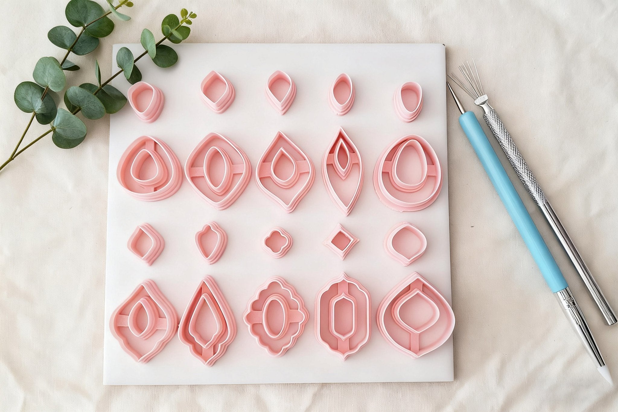 Set of pink polymer clay earring cutters in various shapes arranged on a white board with eucalyptus leaves and sculpting tools.