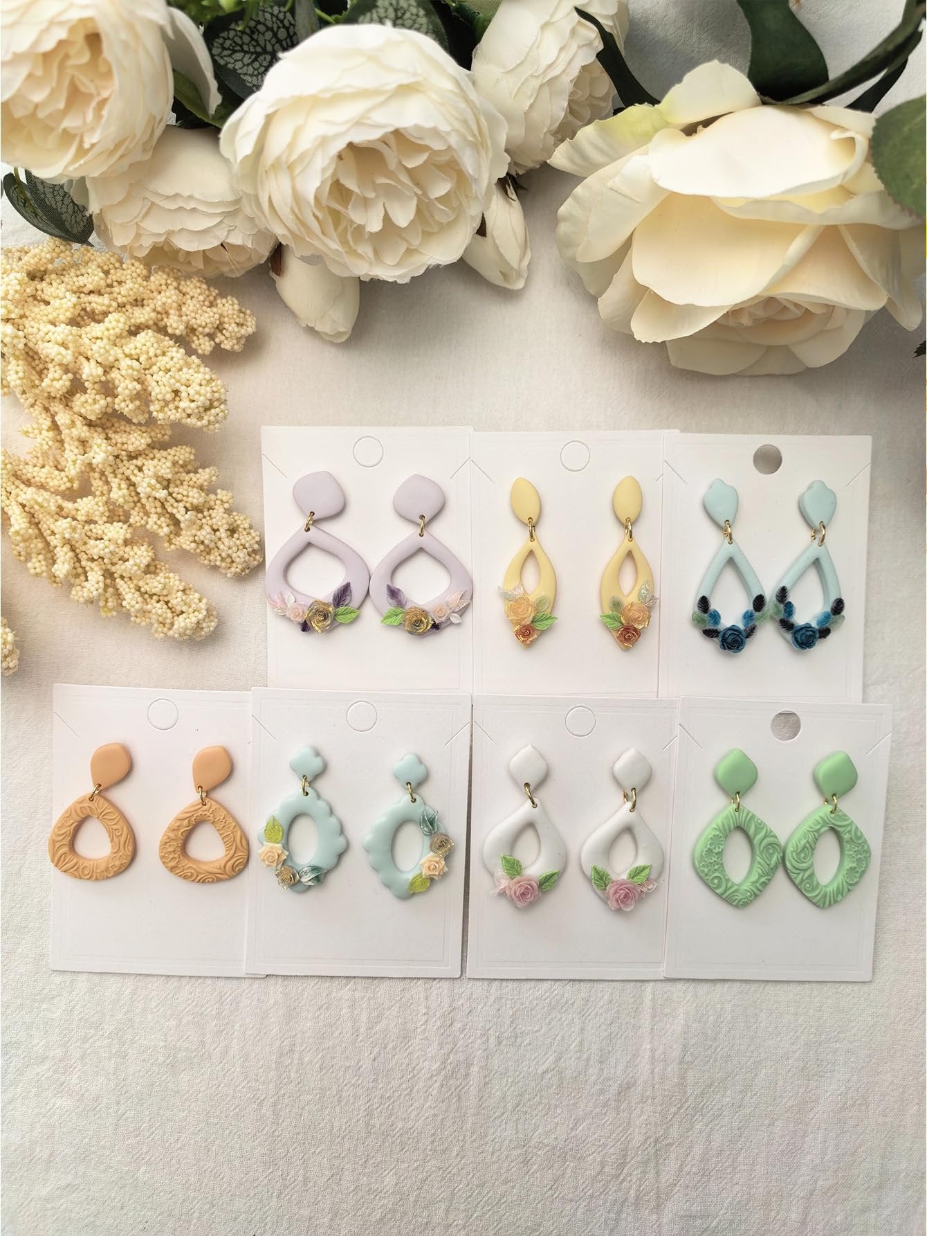 Assortment of polymer clay earrings in various shapes and colors, displayed on white cards with floral embellishments.