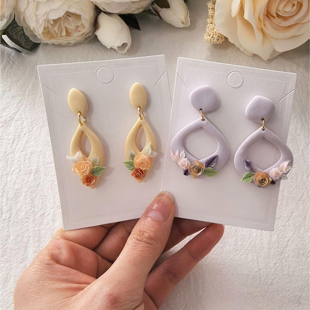 Hand holding two pairs of polymer clay dangle earrings with floral embellishments on white display cards