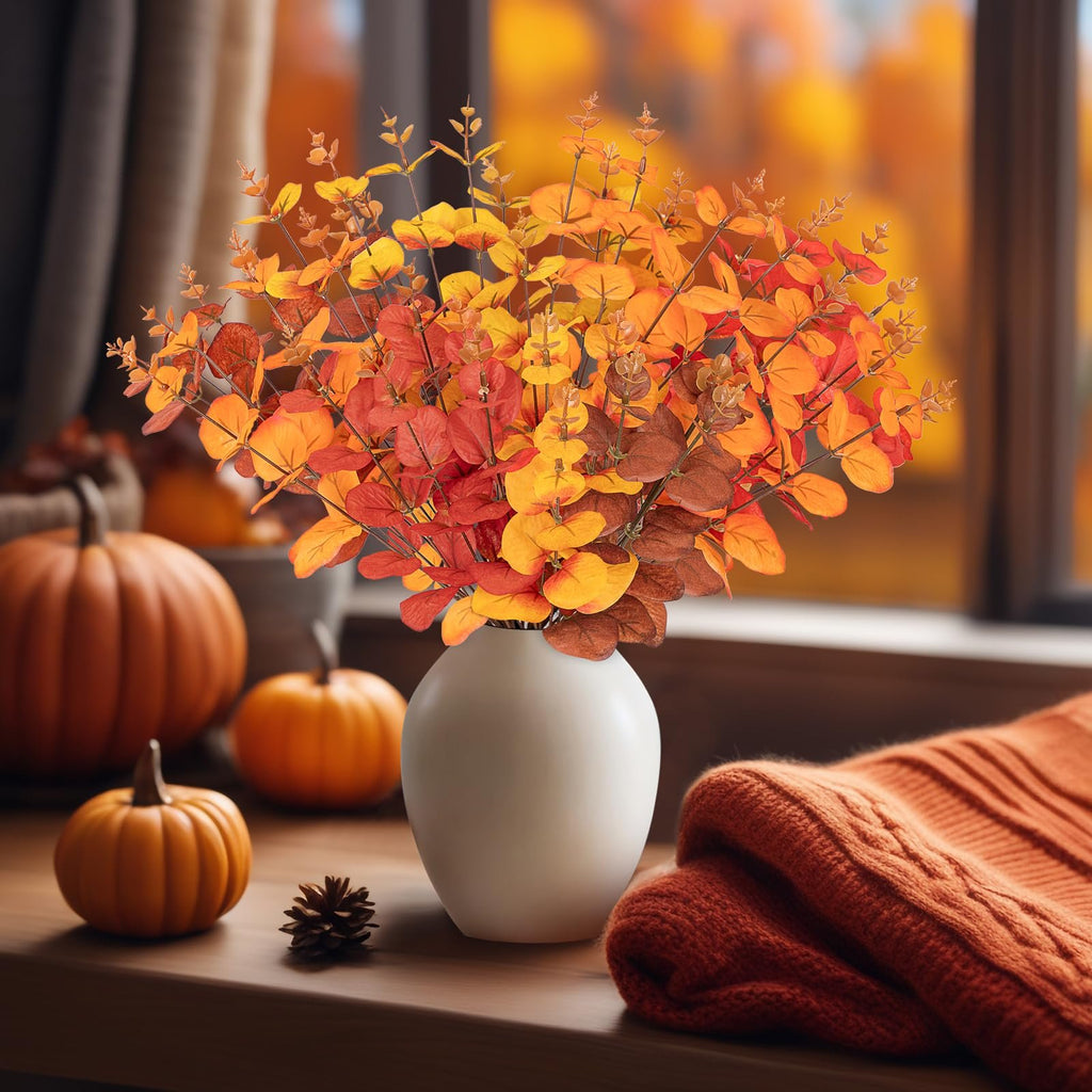 Artificial eucalyptus bouquet in fall colors in a white vase with pumpkins and a sweater on a wooden surface