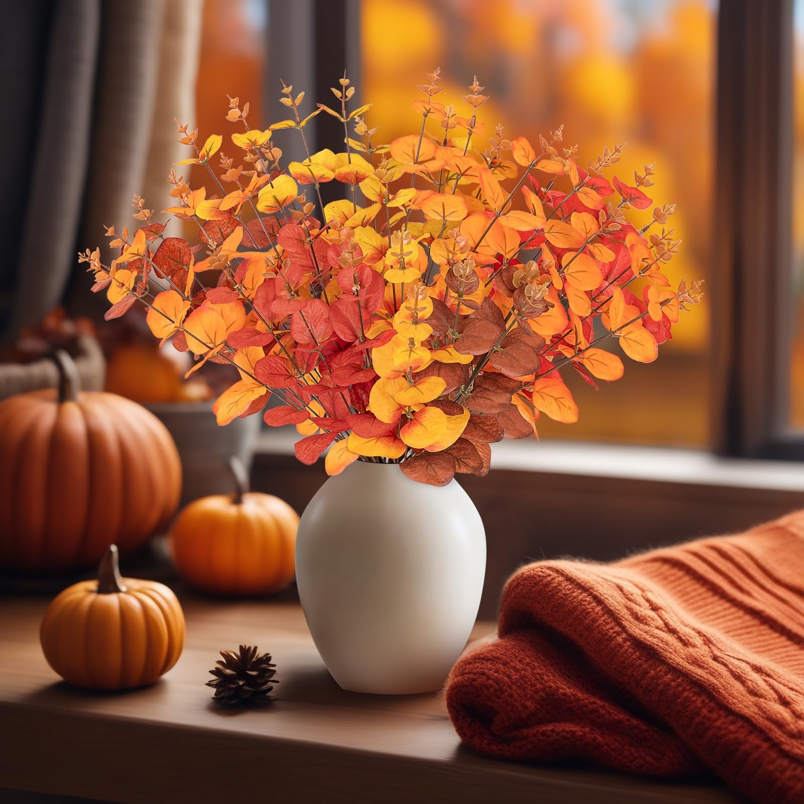 Artificial eucalyptus bouquet in fall colors in a white vase with pumpkins and a sweater on a wooden surface