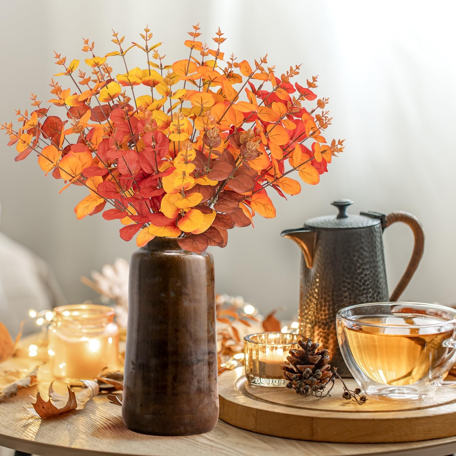 Artificial eucalyptus bouquet in fall colors in a dark vase, with a teapot and tea.