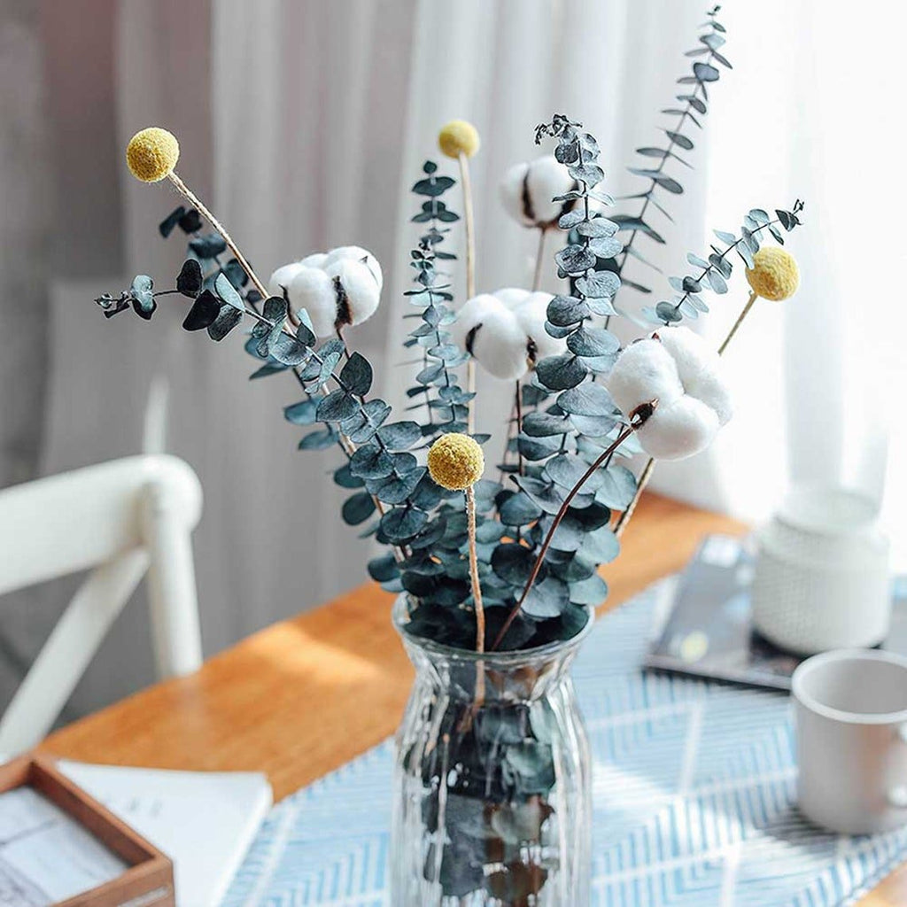 Dried eucalyptus stems with cotton bolls and yellow craspedia in a glass vase on a table.