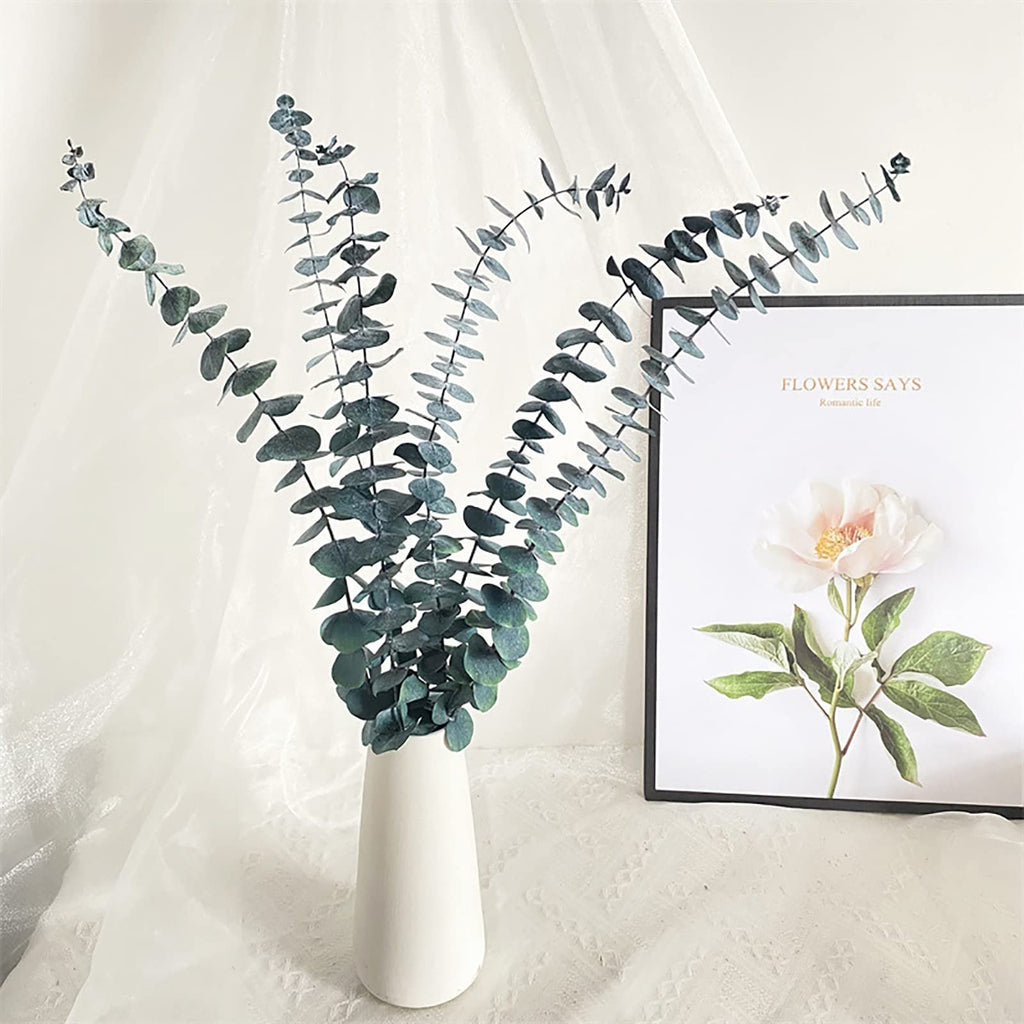 Ten dried eucalyptus stems in a white vase next to a framed floral print with text.