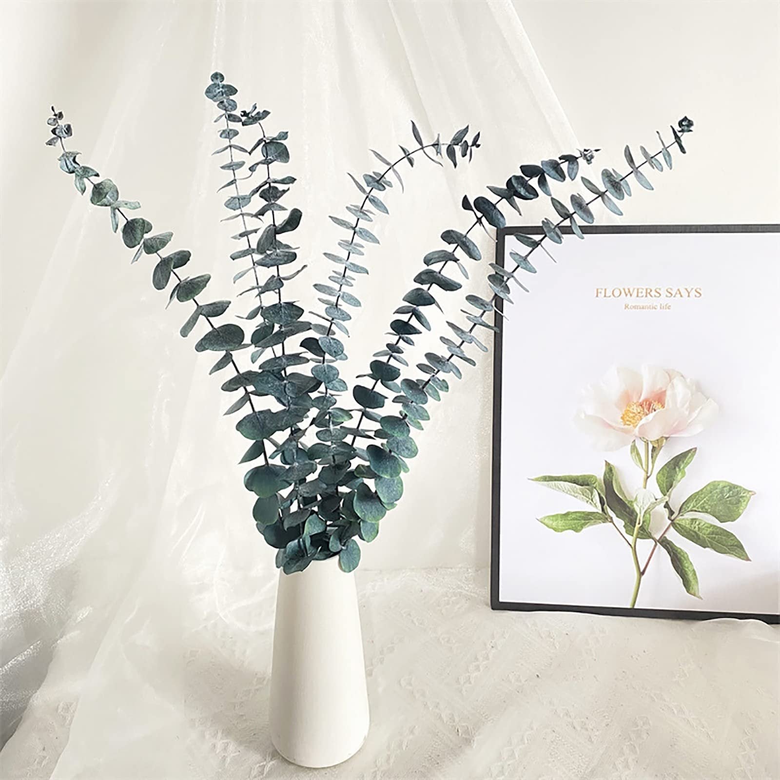 Ten dried eucalyptus stems in a white vase next to a framed floral print with text.