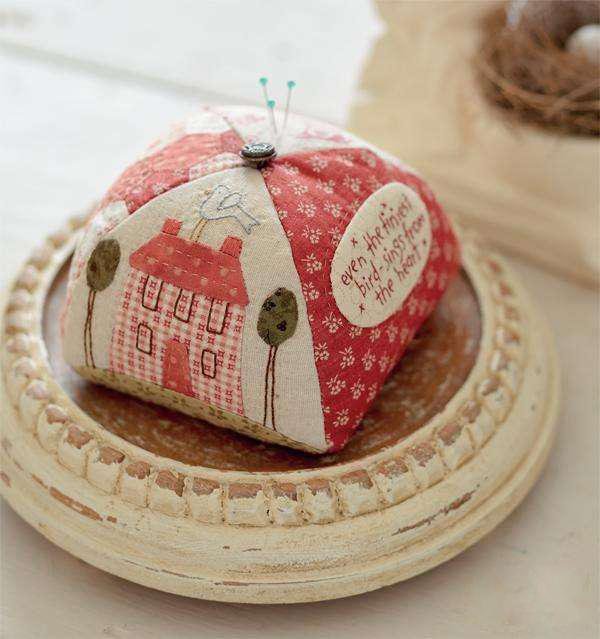 Pincushion shaped like a house with trees, featuring floral and gingham fabric patterns, on a decorative tray.