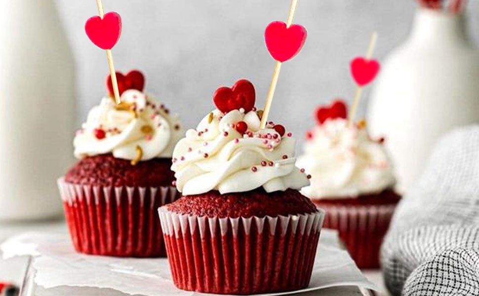 Three red velvet cupcakes topped with white frosting and red heart cocktail picks.