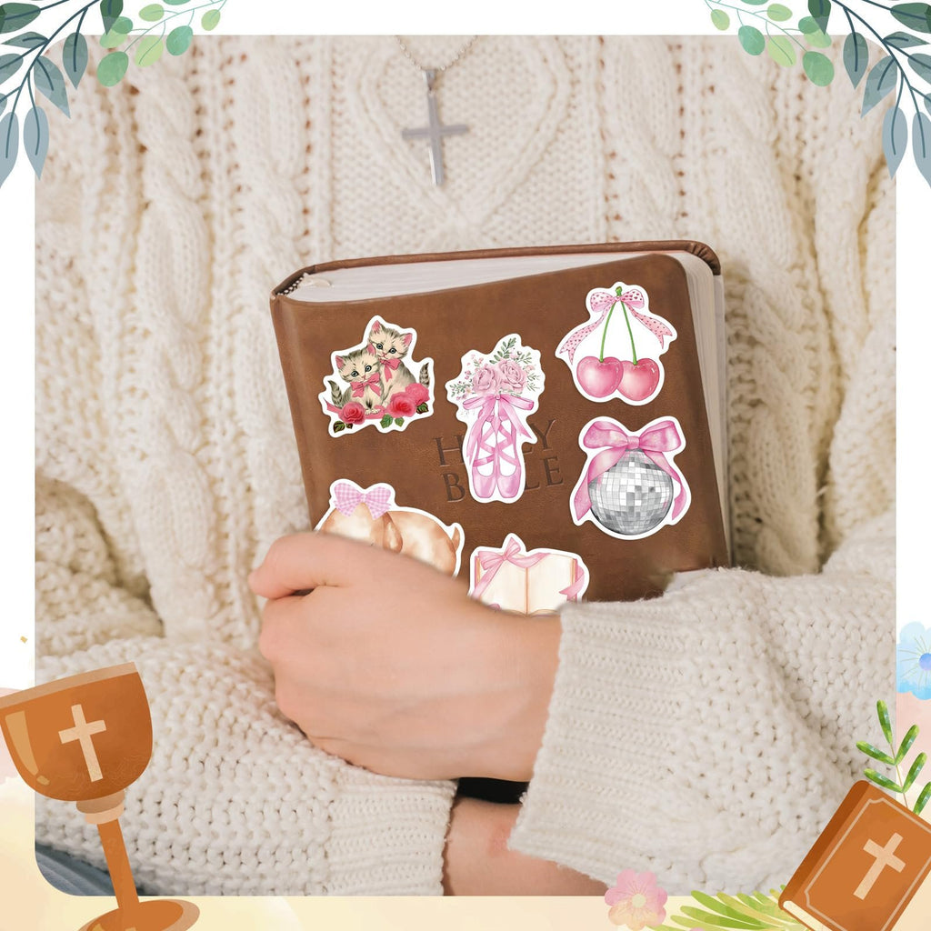 Person holding a Bible with various pink coquette-themed stickers applied, including kittens, ballet slippers, cherries, a disco ball, and a gift.