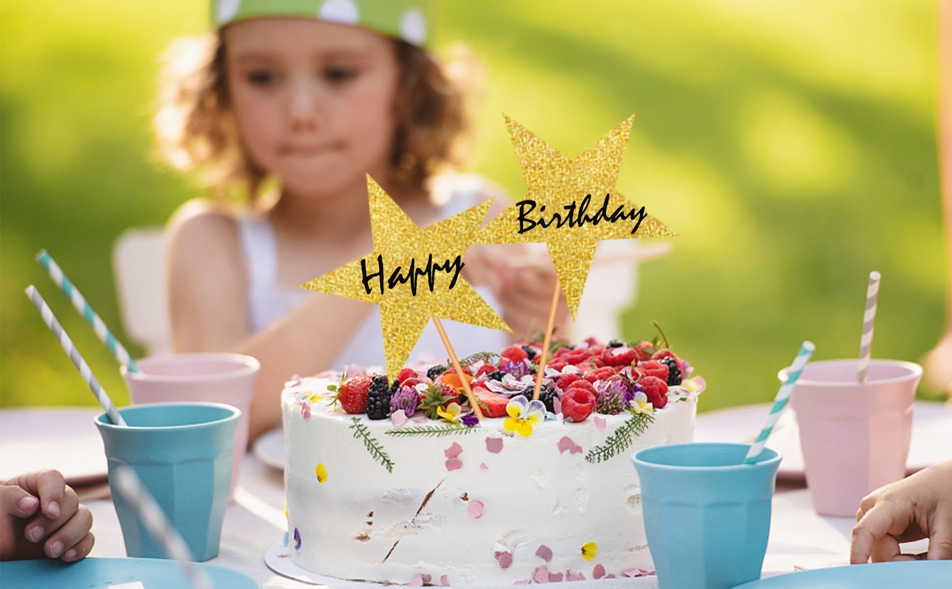 Birthday cake with glitter star toppers that say 'Happy Birthday', at an outdoor party with a child in the background.