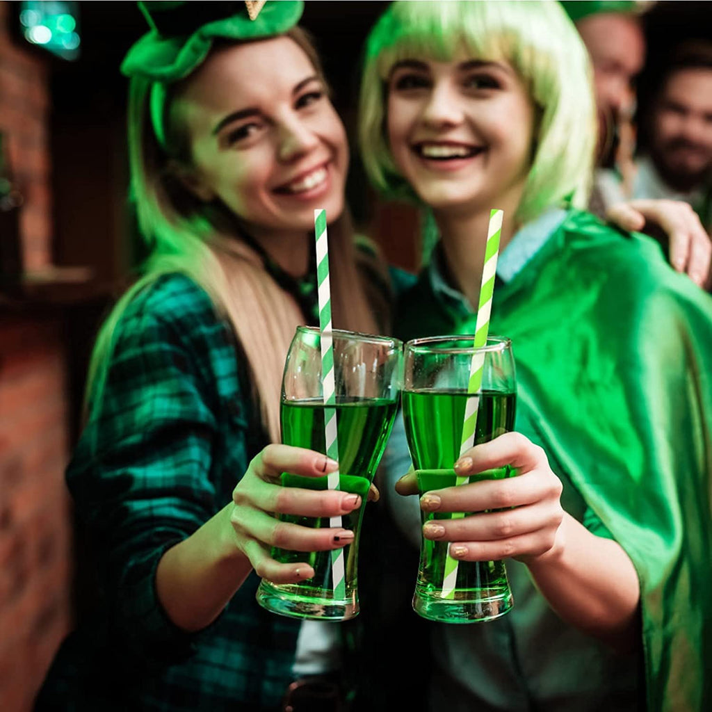 Two smiling women in green hats and clothing clinking glasses of green drinks with striped paper straws.