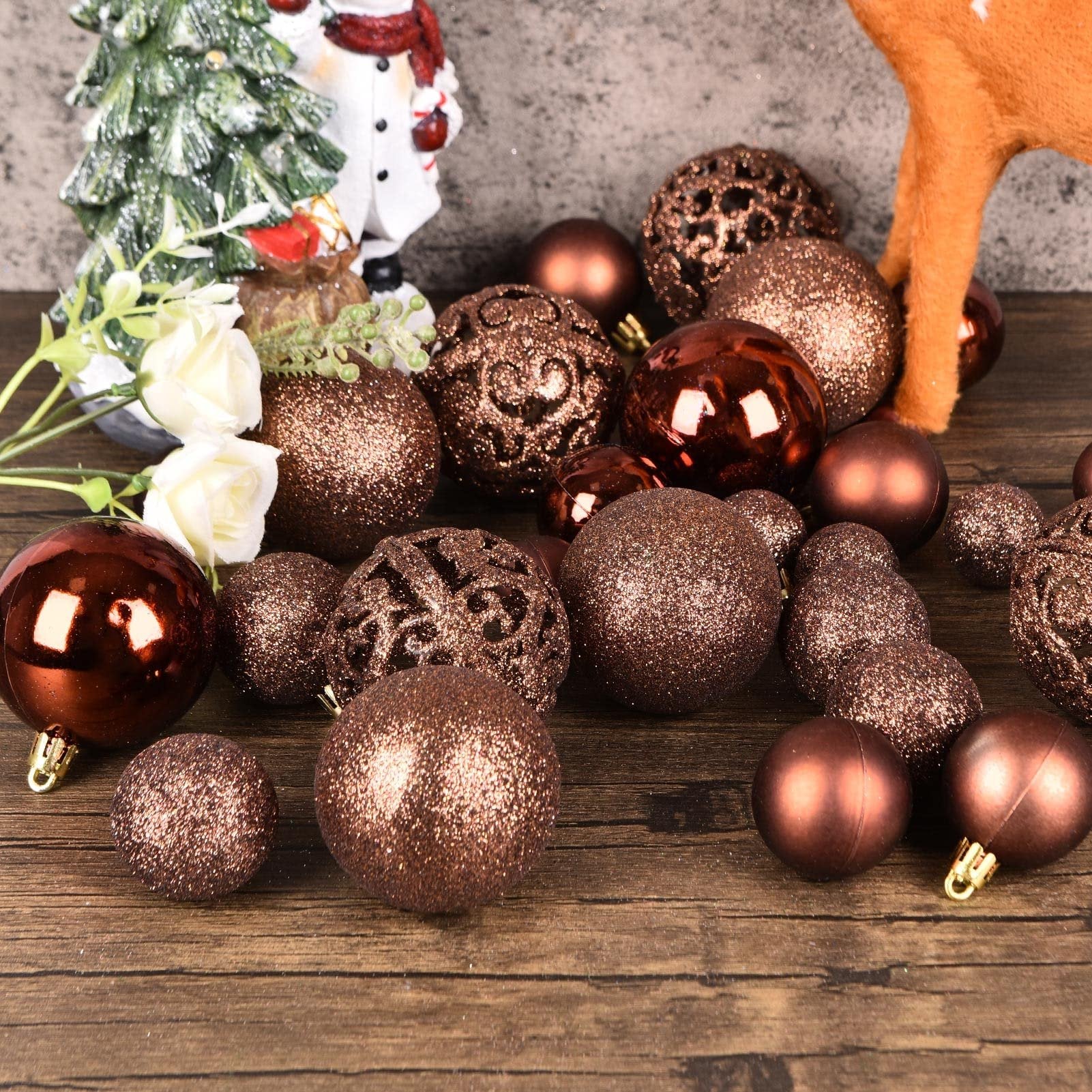 Assortment of brown shatterproof Christmas ball ornaments scattered on a wooden surface with a snowman figurine and white flowers in the background.
