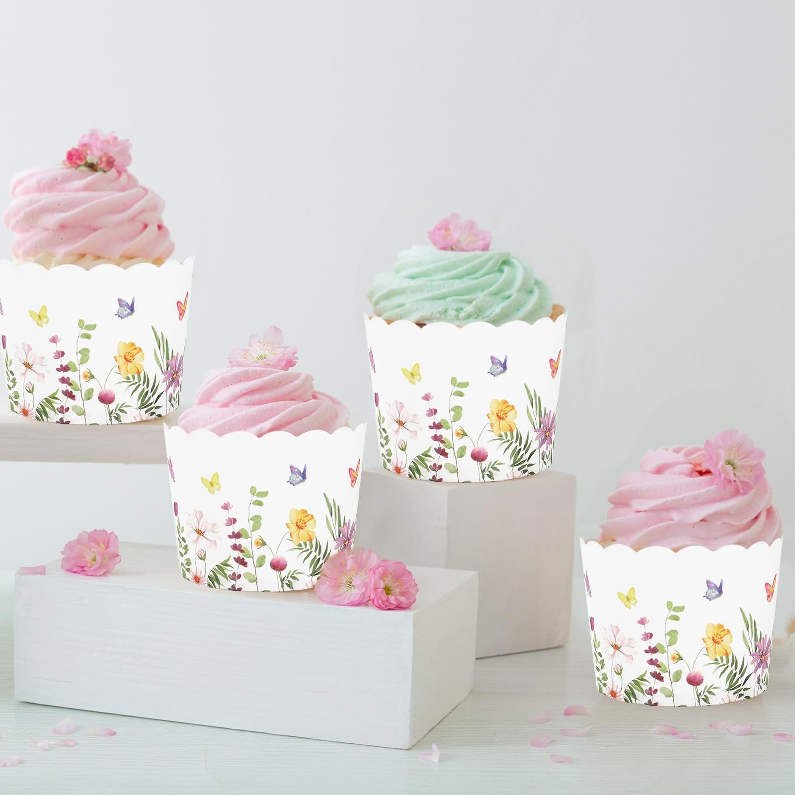 Four decorated cupcakes in wildflower patterned baking cups on white display blocks, with pink flowers and petals scattered around.
