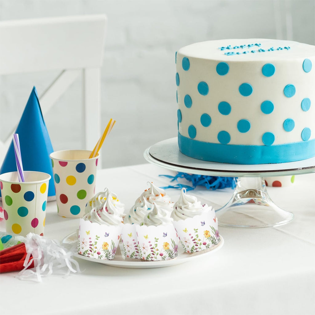 Party table with a polka dot cake, cupcakes in floral wrappers, and colorful cups.