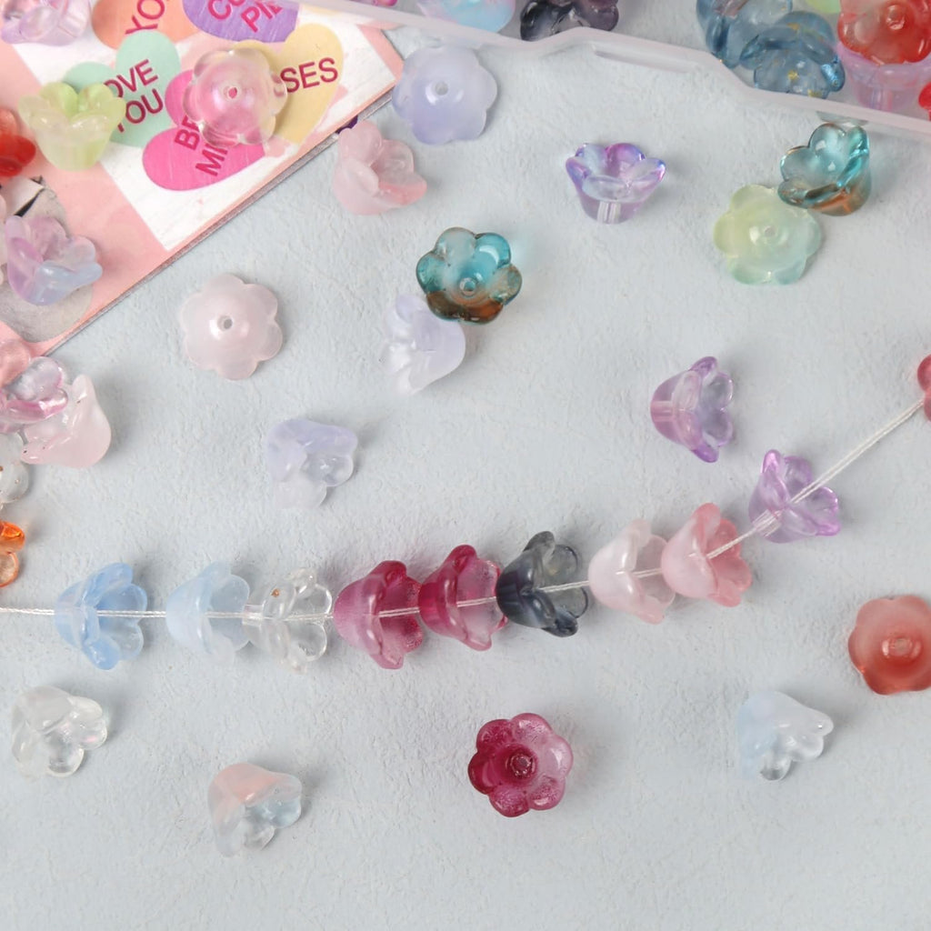 Assorted translucent flower-shaped beads scattered and strung on a white thread, with a pink card featuring heart shapes and text in the background.