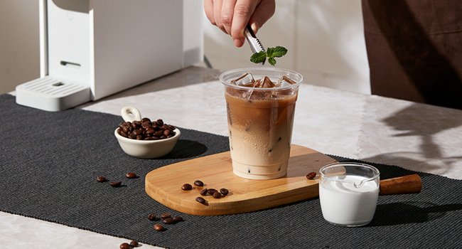 Clear plastic cup with iced coffee and mint garnish, next to coffee beans and milk.