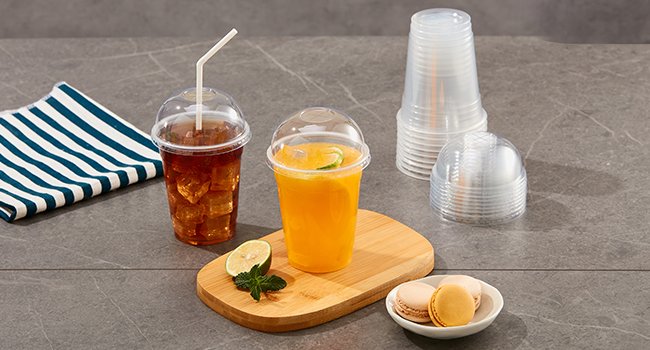 Two clear plastic cups with dome lids, one filled with iced tea and a straw, the other with orange juice and lime slices, next to macarons and a stack of cups.