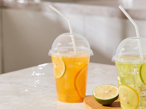 Two clear plastic cups with dome lids and straws, one filled with orange drink and citrus slices, the other with a green drink and lime slices.