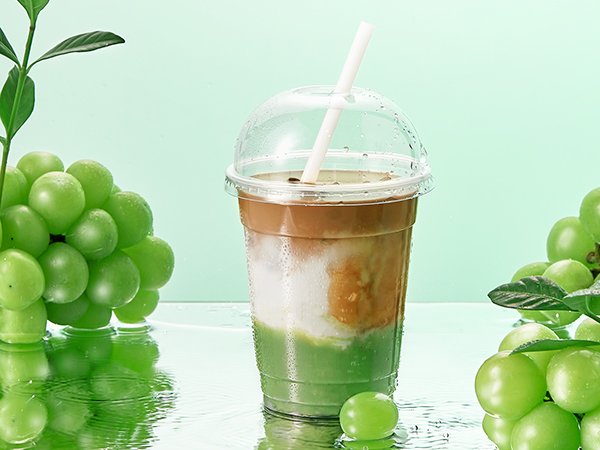 Clear plastic cup with dome lid and straw filled with layered green and brown drink, surrounded by green grapes and reflections.