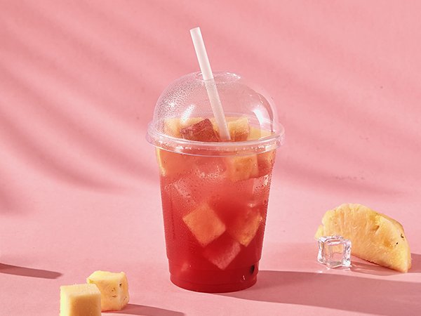 Clear plastic cup with dome lid filled with red drink and fruit chunks, with pineapple slices and ice cubes on pink background.