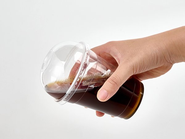Hand holding a clear plastic cup filled with dark liquid and foam, with a dome lid on top.