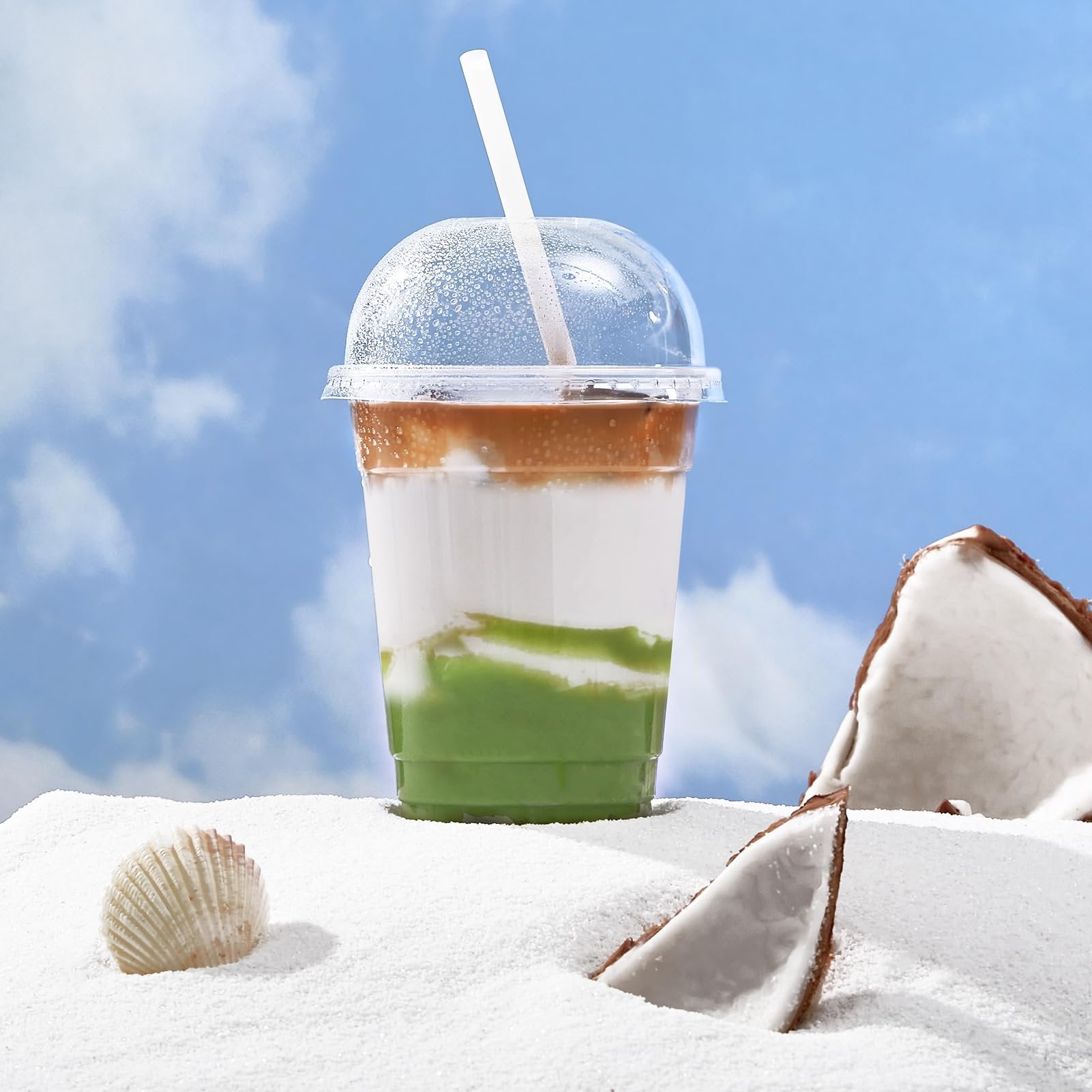 Clear plastic cup with dome lid filled with layered drink, on white sand with coconut pieces and a seashell.