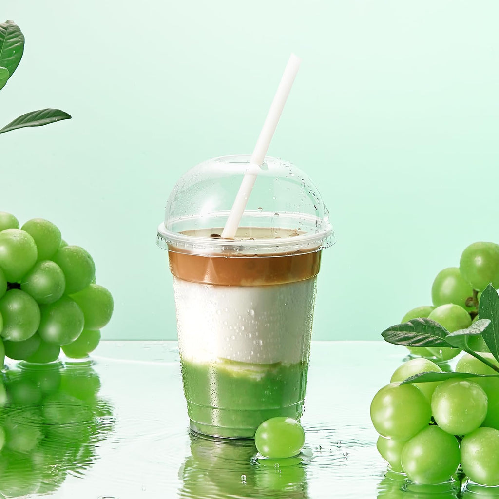 Clear plastic cup with dome lid filled with layered green and white drink topped with coffee, with a straw, surrounded by green grapes and water droplets.