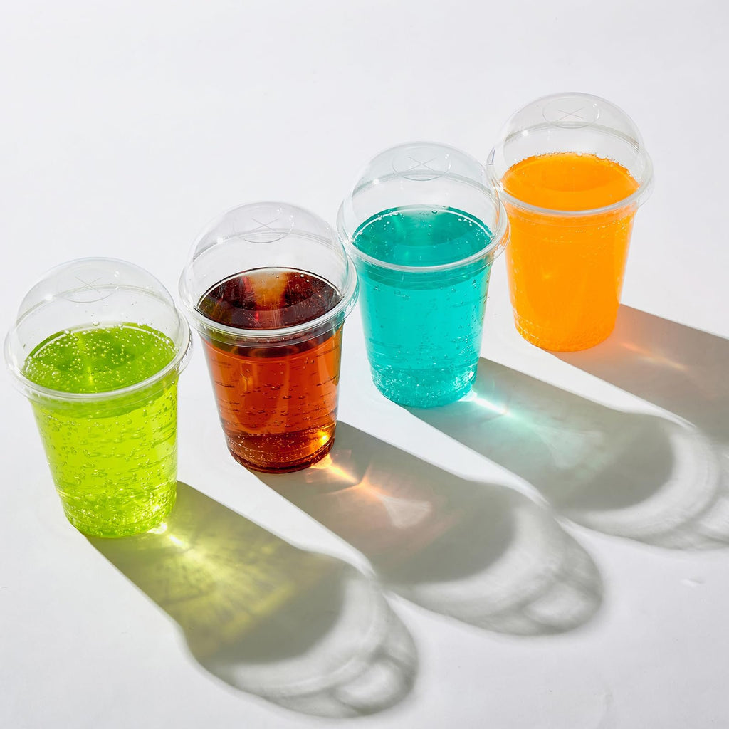 Four clear plastic cups with dome lids filled with colorful beverages, arranged in a row on a white surface.
