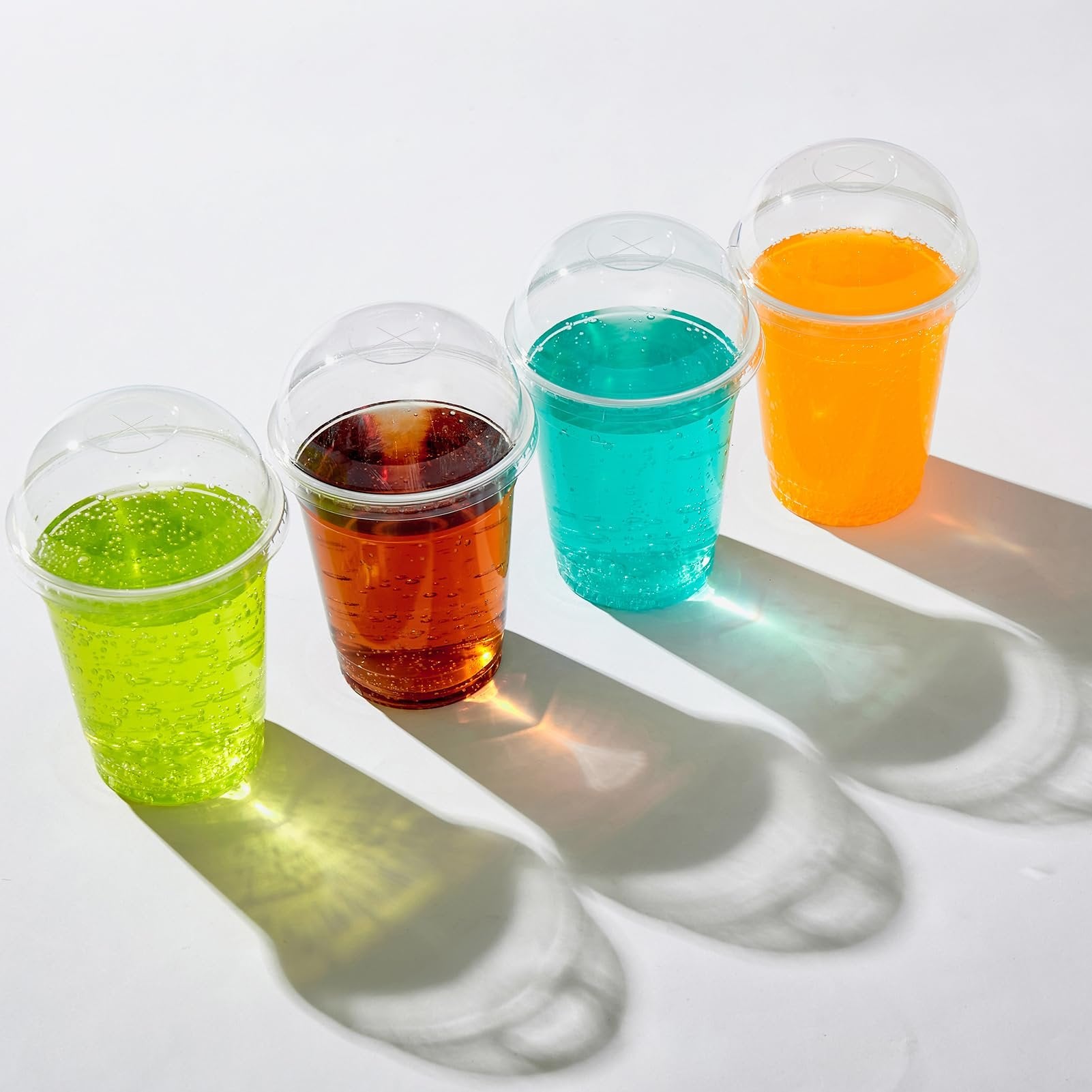 Four clear plastic cups with dome lids filled with colorful beverages, arranged in a row on a white surface.