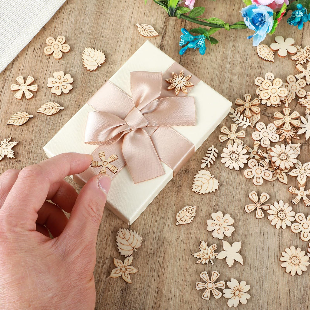 Hand holding a wooden flower cutout over a gift box surrounded by assorted wooden flower and leaf craft ornaments on a wooden surface.