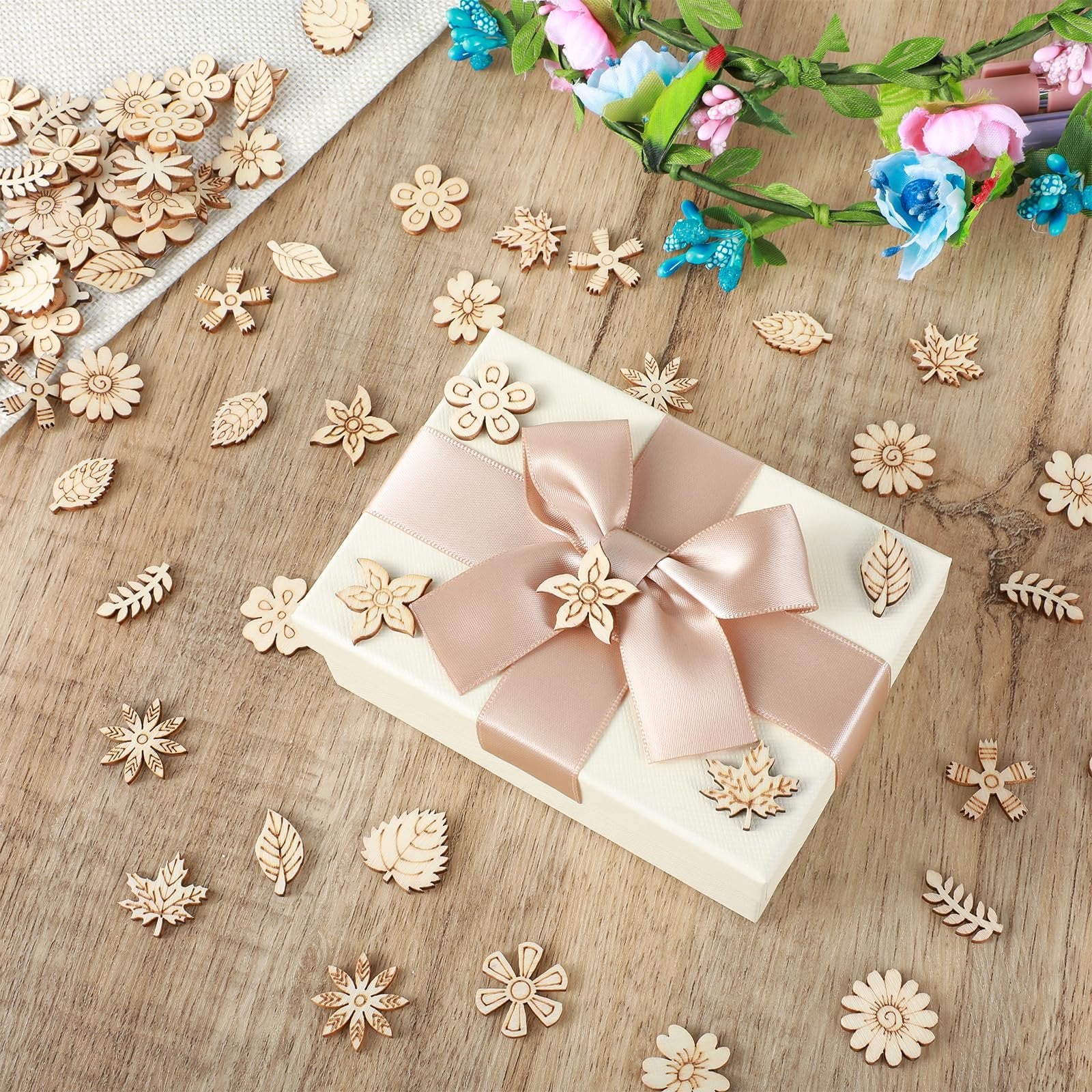 Assortment of 20mm unfinished wooden flower and leaf craft cutouts scattered around a gift box with a pink ribbon.