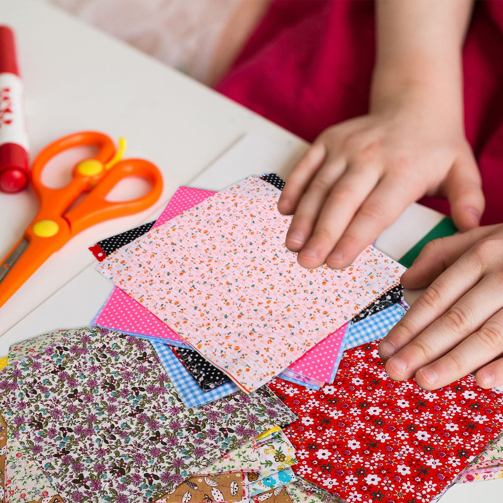 Stack of colorful floral printed cotton fabric squares with scissors and a glue stick on a white table.