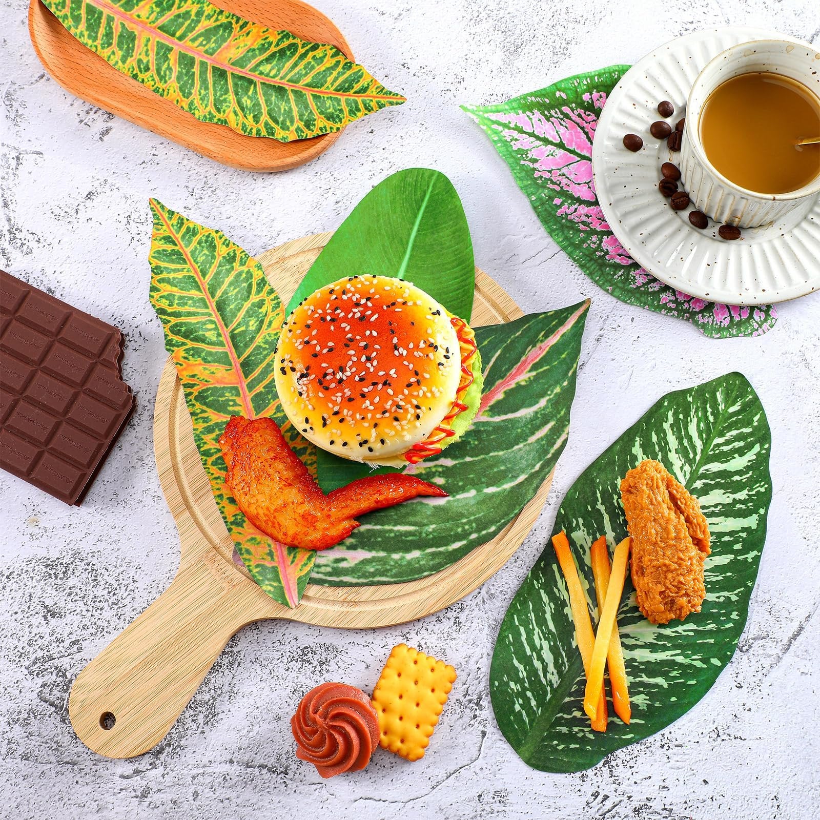 Tropical leaf parchment paper serving as placemats for food items like a burger, chicken wing, fries, chocolate, and coffee.
