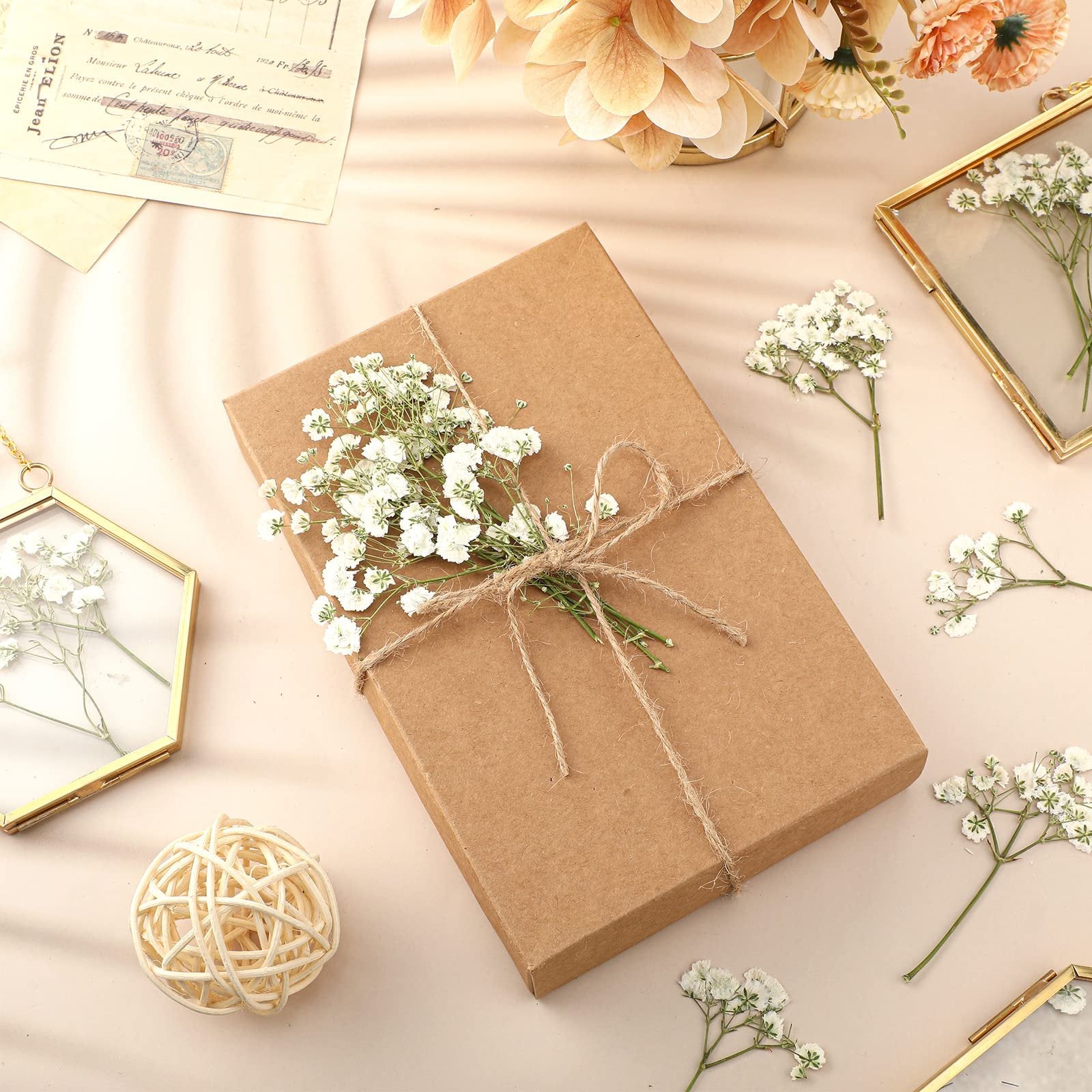 Brown gift box tied with twine and decorated with dried baby's breath flowers, surrounded by more flowers and decorative frames.