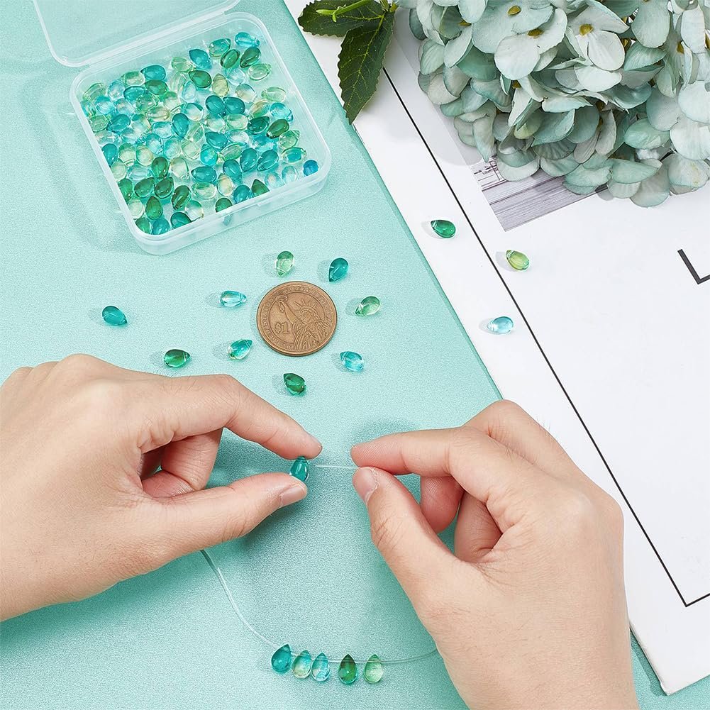 Hands stringing teardrop-shaped glass beads in blue and green hues, with a container of beads and a coin for scale.