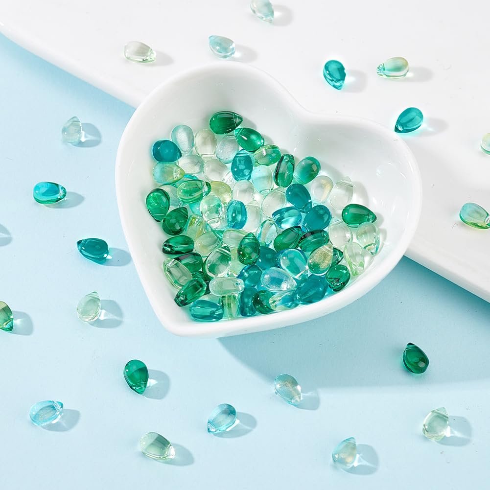 Teardrop glass beads in blue and green scattered around a white heart-shaped dish filled with beads.